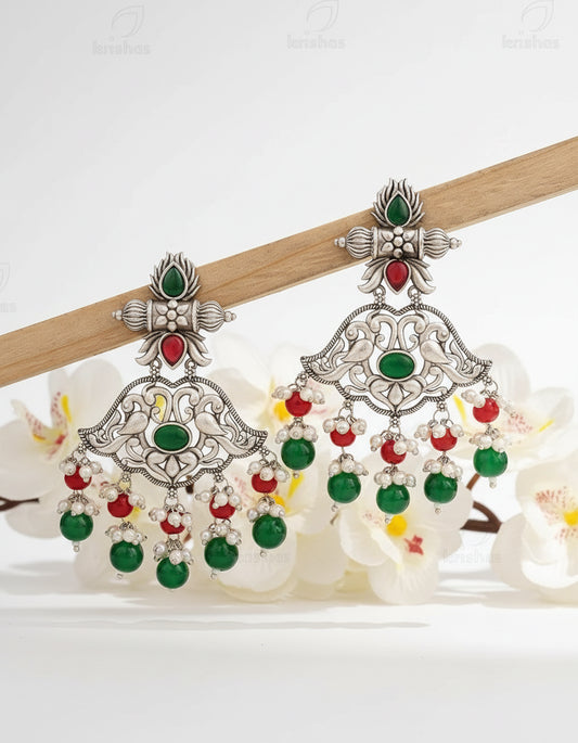 Ornate silver earrings with green and red stones on a wooden stick against a brown background
