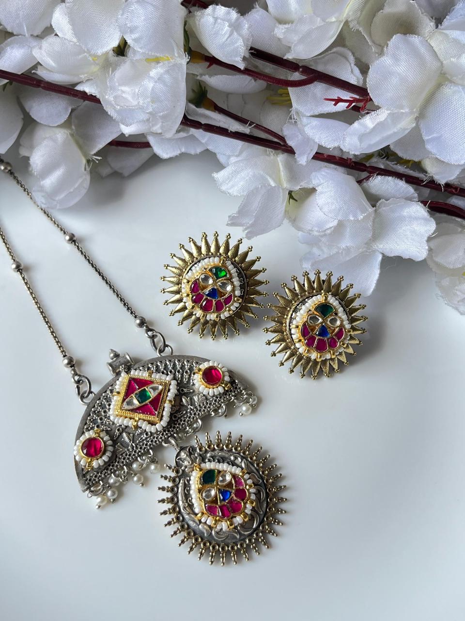 Ornate jewelry set with red and green gemstones on a white surface with white flowers in the background