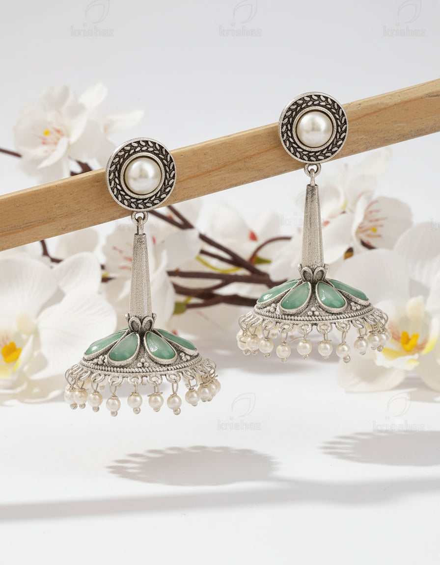 Pair of silver earrings with green and pearl accents on a wooden stick against a brown background