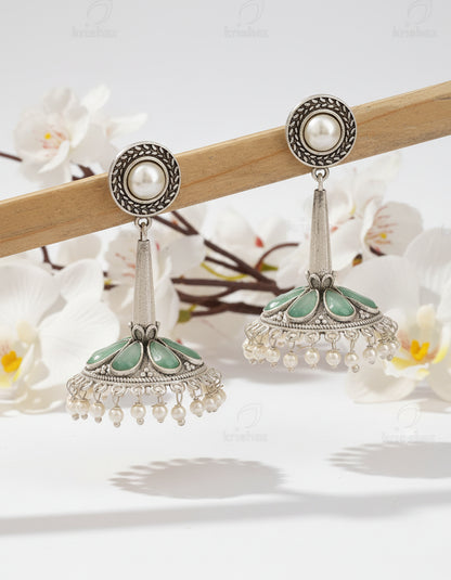 Pair of silver earrings with green and pearl accents on a wooden stick against a brown background