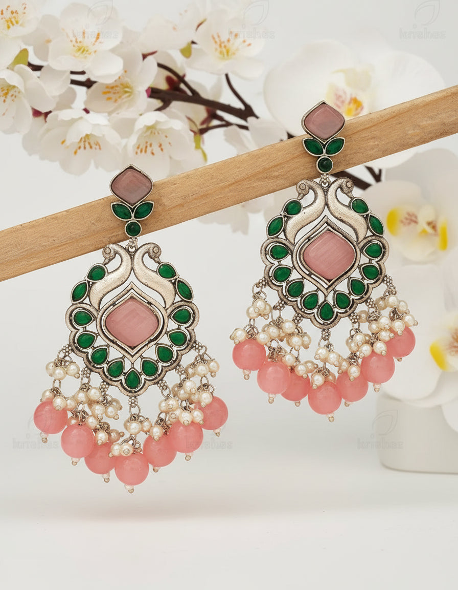 Ornate earrings with pink stones and pearl-like beads on a wooden stick against a brown background