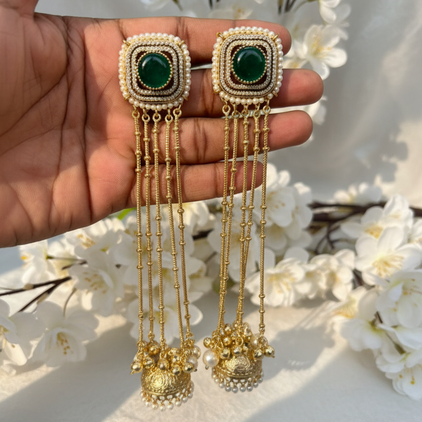 Gold earrings with green gemstones held in a hand against a white floral background