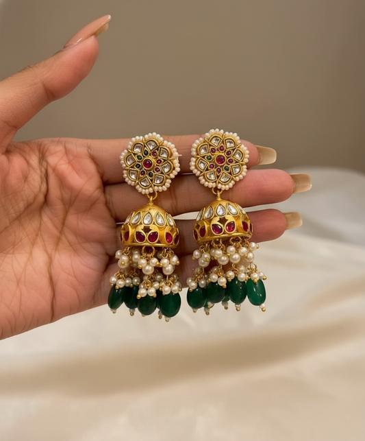 Ornate earrings with pearls and gemstones held by a hand against a neutral background