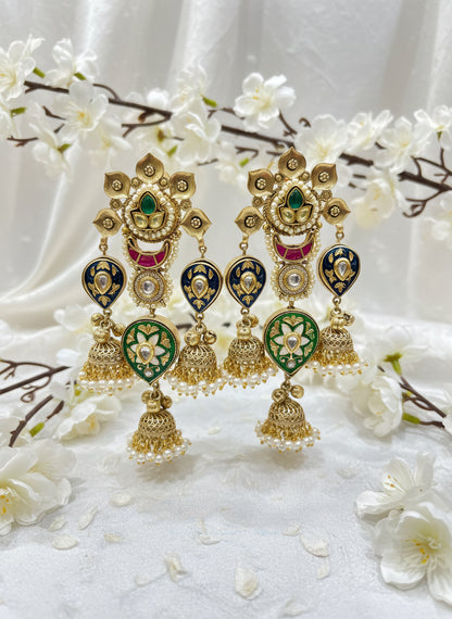 Gold and green decorative earrings held by a hand with a blurred indoor background.