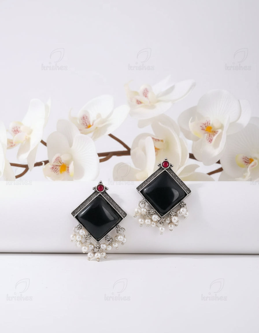 Pair of earrings with black square stones, red gemstones, and pearl details on a white surface.